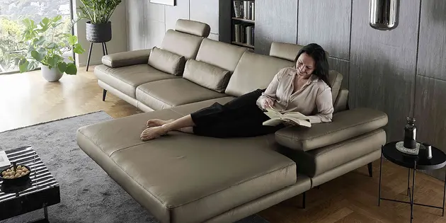 Modernes Wohnzimmer mit großem olivfarbenem Ledersofa und breiter Recamiere auf schwarzen Metallfüßen. Frau sitzt entspannt mit Smartphone, graue Wand mit Bücherregal und Bild, dunkler Teppich und eleganter Couchtisch im Vordergrund.
